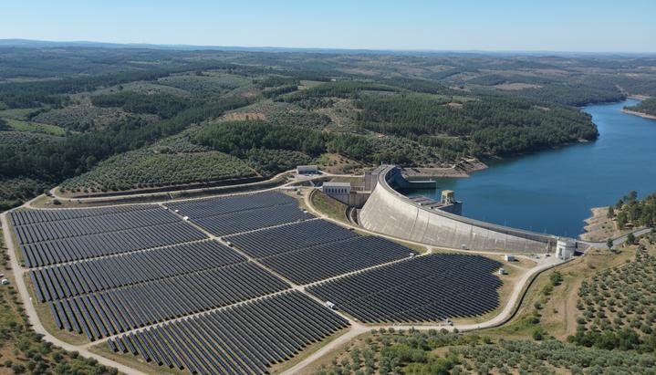 Солнечная электростанция с рядами фотоэлектрических панелей на фоне бетонной плотины гидроэлектростанции на реке среди зеленых холмов.
